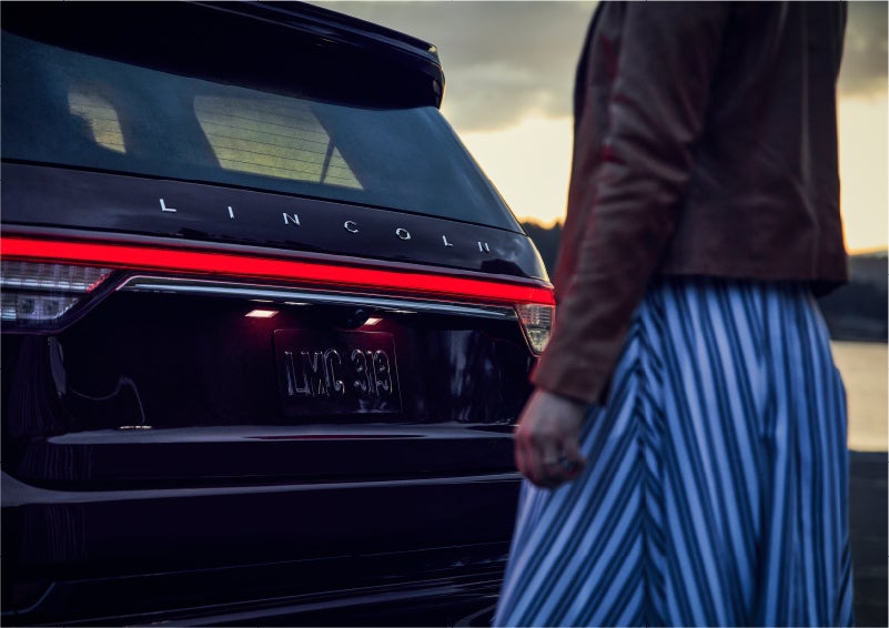 A person is shown near the rear of a 2023 Lincoln Aviator® SUV as the Lincoln Embrace illuminates the rear lights | Magic City Lincoln in Roanoke VA